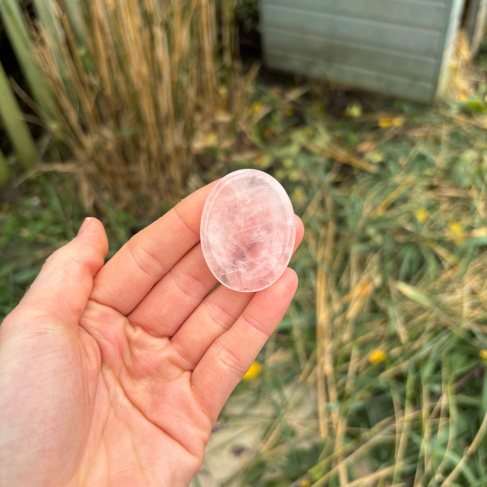 A hand presenting the polished rose quartz worry stone at a distance over natural outdoor grass, demonstrating its physical 4cm sizing scale as a comforting pocket companion to help melt away daily stress.
