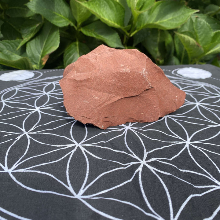 An alternative high-angle view looking down at the rough-cut red jasper crystal resting on a black sacred geometry fabric mat. The natural sunlight captures its smooth yet unpolished matte surfaces, bringing a reassuring sense of warmth, balance, and protection to any space.
