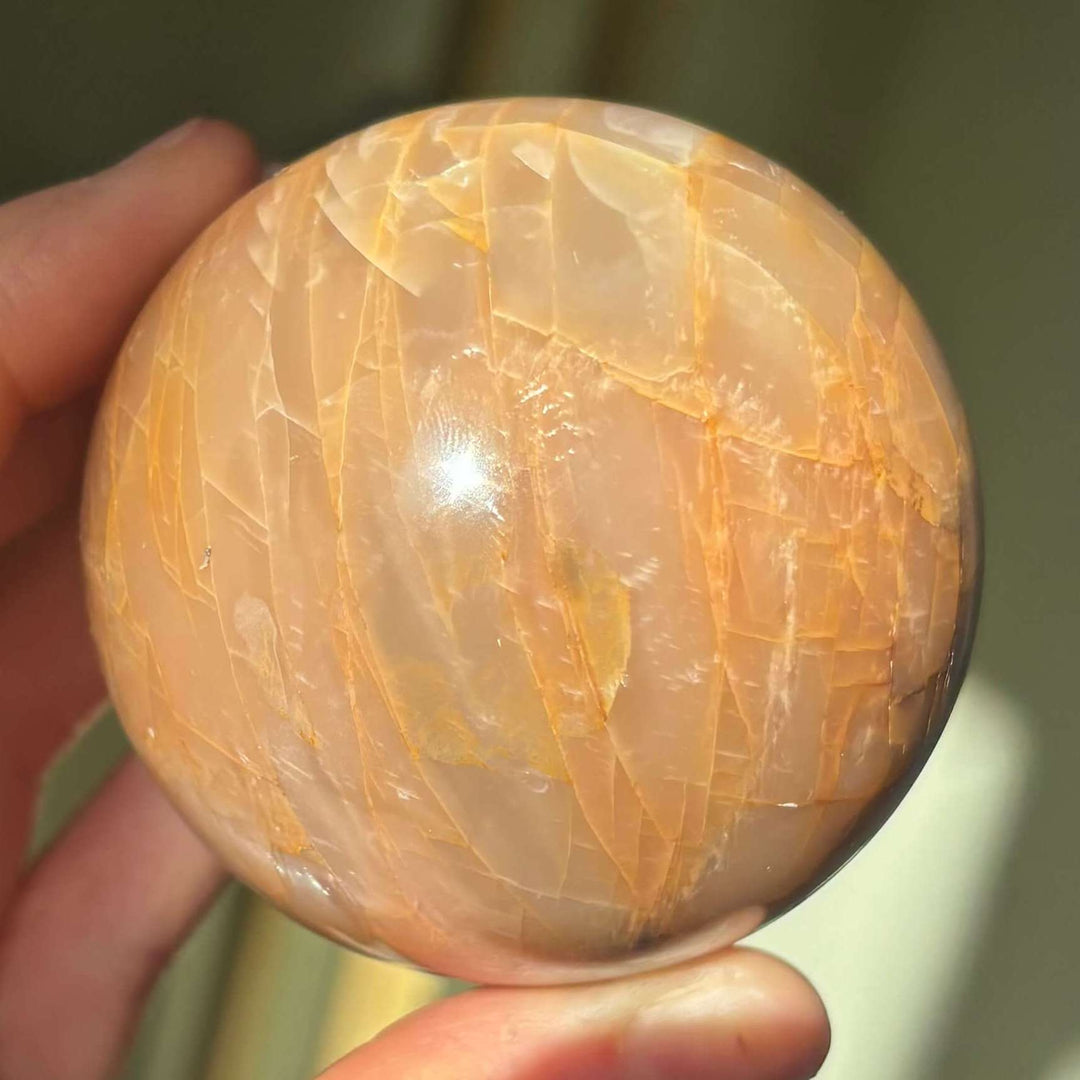 A hand gently balancing the 357-gram Peach Moonstone gemstone ball under daylight, highlighting the natural shimmer and glow ideal for sacral chakra alignment.