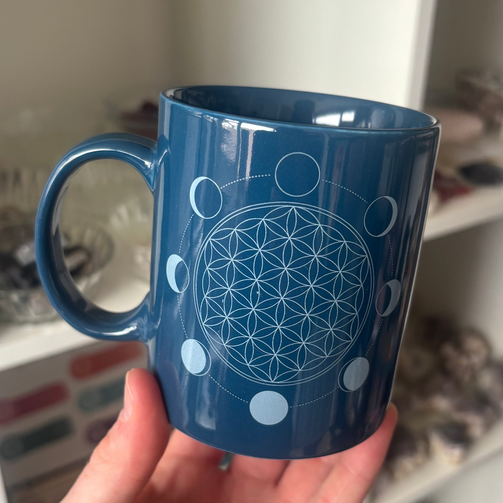 An eye-level shot showing the Moon Phase Flower of Life ceramic mug held carefully in a hand. Displayed against a blurred indoor background of shop shelves, the smooth glossy finish and cosmic alignment details provide a cosy, grounding everyday vibe.