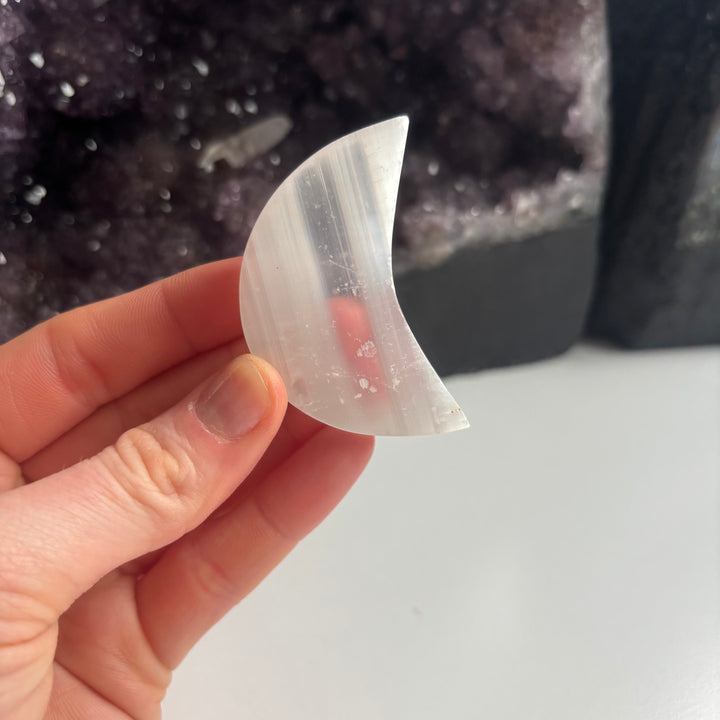 A hand holding the thin side profile of a satin spar selenite moon carving from the imperfect collection, illustrating the smooth curved edges designed to invite a soft frequency of calm serenity.