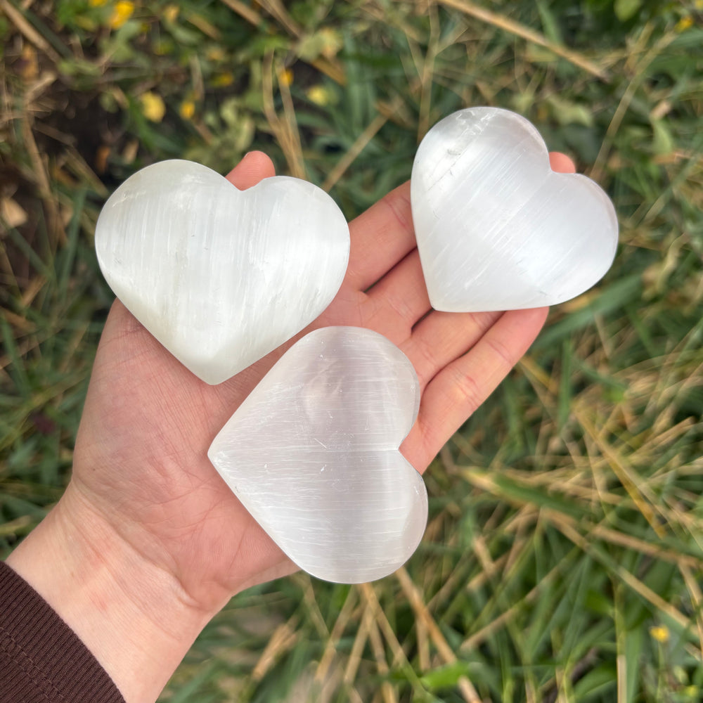 A hand holding three polished satin spar selenite pocket hearts at a wider distance outdoors over green garden grass, demonstrating the physical sizing scale of these comforting celestial tokens designed to ease daily stress.