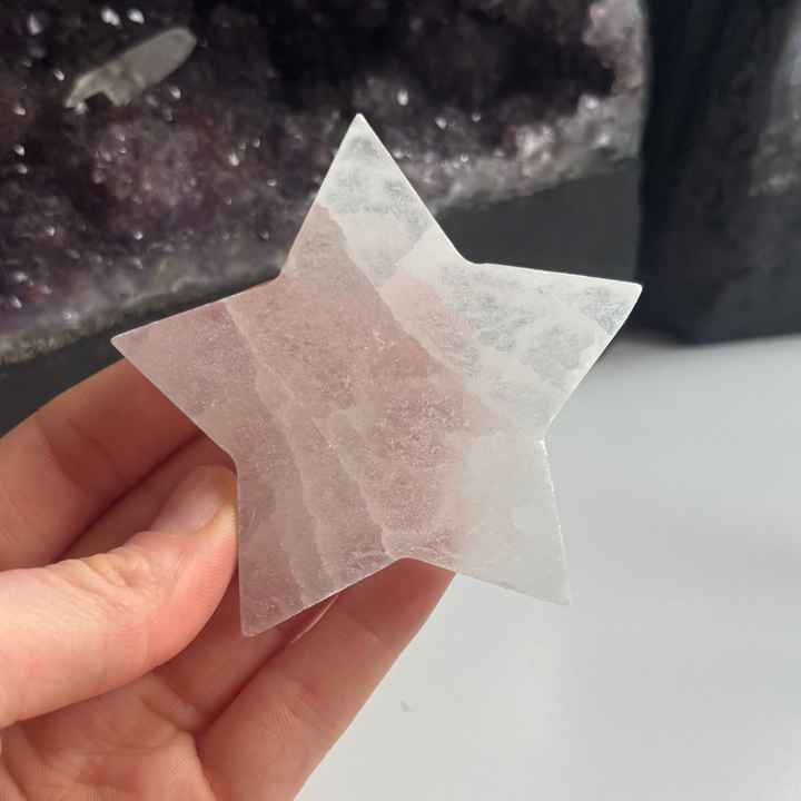 A hand showing the detailed front face of the satin spar selenite star carving indoors, demonstrating how the reflective mineral layers catch light to serve as a visual anchor during evening relaxation routines.
