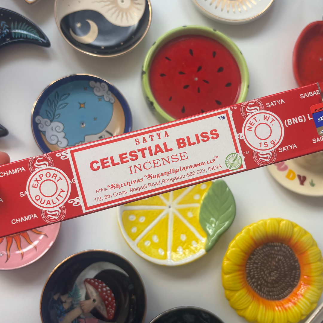 A direct overhead shot showcasing a standard 15g rectangular cherry-red box of authentic Satya Celestial Bliss incense sticks held diagonally across the frame. The retail packaging features a clean white label with bold red "Celestial Bliss Incense" lettering, resting above a flat lay collection of colourful ceramic trinket dishes including a yellow lemon slice, a red watermelon design, and a sunflower bowl.