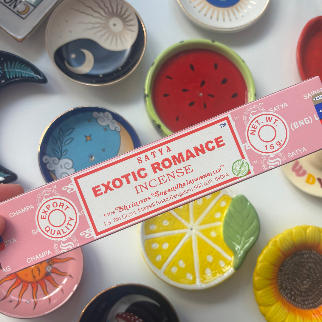 A direct overhead shot showcasing a standard 15g rectangular rose-pink box of authentic Satya Exotic Romance incense sticks held diagonally across the frame. The retail packaging features a clean white label with bold red "Exotic Romance Incense" lettering, resting above a flat lay collection of colourful ceramic trinket dishes including a yellow lemon slice, a red watermelon design, and a sunflower bowl.