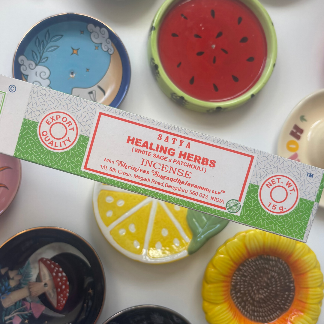 A direct overhead shot showcasing a standard 15g rectangular light green and grey patterned box of authentic Satya Healing Herbs incense sticks held diagonally across the frame. The retail packaging features a clean white central accent label with bold red "Healing Herbs Incense" lettering, resting above a flat lay collection of colourful ceramic trinket dishes including a yellow lemon slice, a red watermelon design, and a sunflower bowl.