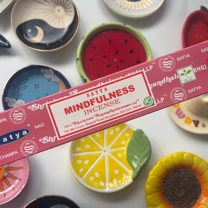 A direct overhead shot showcasing a standard 15g rectangular berry-pink box of authentic Satya Mindfulness incense sticks held diagonally across the frame. The retail packaging features a clean white label with bold red "Mindfulness Incense" lettering, resting above a flat lay collection of colourful ceramic trinket dishes including a yellow lemon slice, a red watermelon design, and a sunflower bowl.