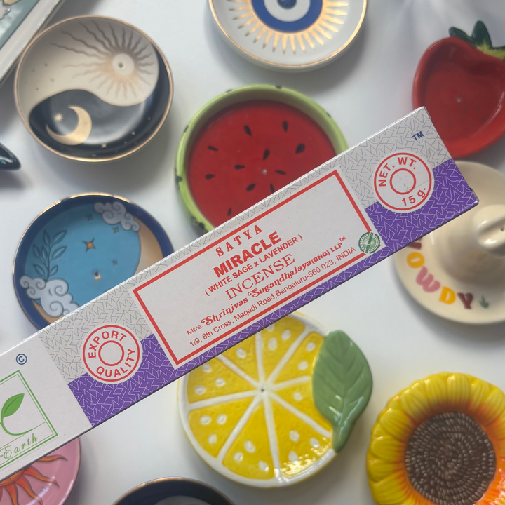 A direct overhead shot showcasing a standard 15g rectangular light purple and white box of authentic Satya Miracle incense sticks held diagonally across the frame. The retail packaging features a clean white accent label with bold red "Miracle Incense" lettering, resting above a flat lay collection of colourful ceramic trinket dishes including a yellow lemon slice, a red watermelon design, and a sunflower bowl.