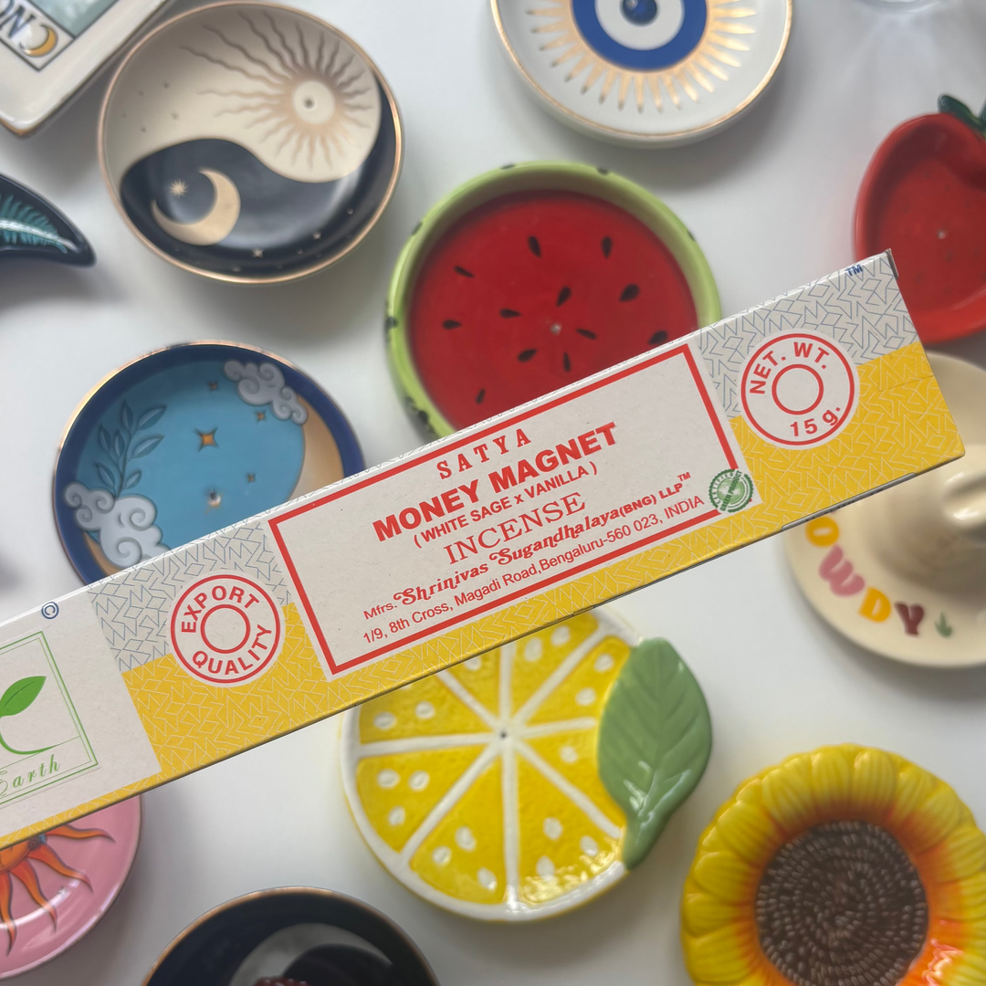 A direct overhead shot showcasing a standard 15g rectangular bright yellow box of authentic Satya Money Magnet incense sticks held diagonally across the frame. The retail packaging features a white accent label with bold red "Money Magnet Incense" lettering, resting above a flat lay collection of colorful ceramic trinket dishes including a yellow lemon slice, a red watermelon design, and a sunflower bowl.
