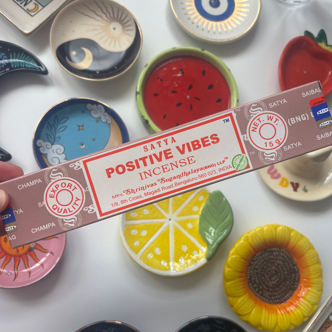 A direct overhead shot showcasing a standard 15g rectangular light brown box of authentic Satya Positive Vibes incense sticks held diagonally across the frame. The retail packaging features bold red "Positive Vibes Incense" lettering and traditional circular quality marks, resting above a flat lay collection of colorful ceramic trinket dishes including a yellow lemon slice, a red watermelon ring, and a sunflower bowl.