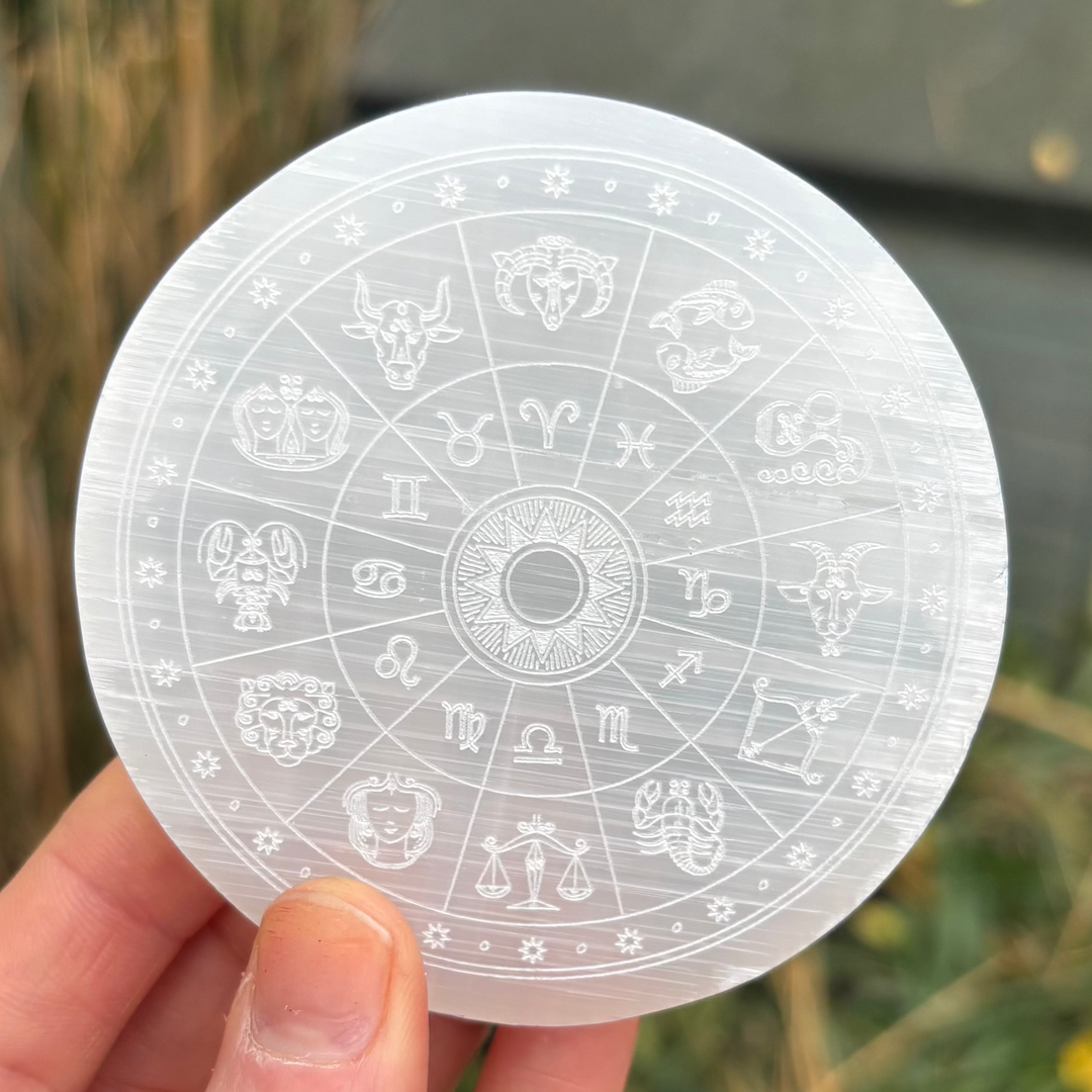 A close-up view of a hand holding a polished selenite charging plate disc outdoors, showcasing the detailed laser-engraved horoscope zodiac wheel symbols and fine natural fibrous lines catching daylight for Crown Chakra alignment.