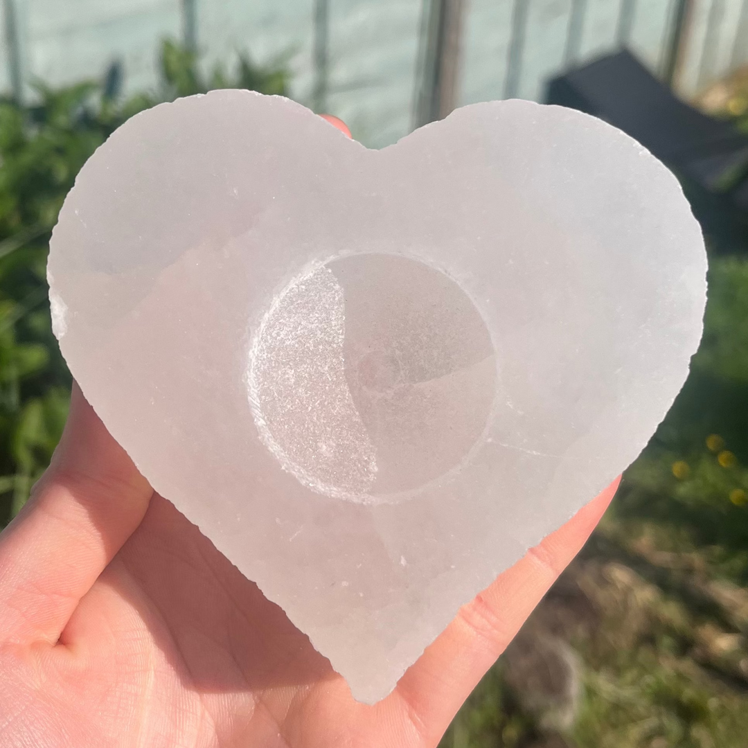 A close-up view of an open hand supporting a carved selenite love heart tea-light holder outdoors, showcasing its ice-like shimmer, circular candle indent, and natural fibrous textures catching bright daylight for Crown and Heart Chakra alignment.