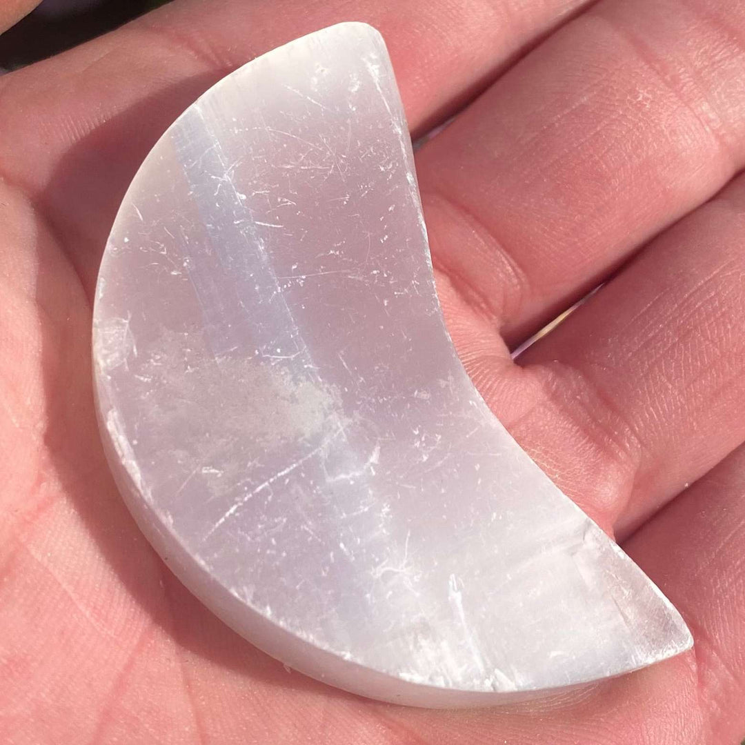 A polished Selenite moon carving resting in an open hand, showcasing its smooth crescent shape, pearly white surface, and natural fibrous lines under bright sunlight.