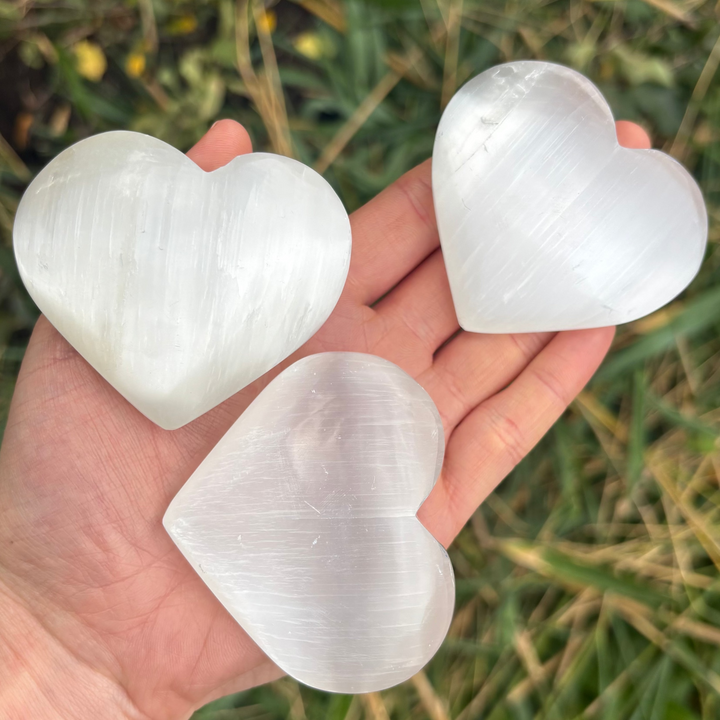 An open hand cradling three polished selenite puff heart crystal carvings outdoors, showcasing their smooth 3D rounded contours, bright white composition, and cat&