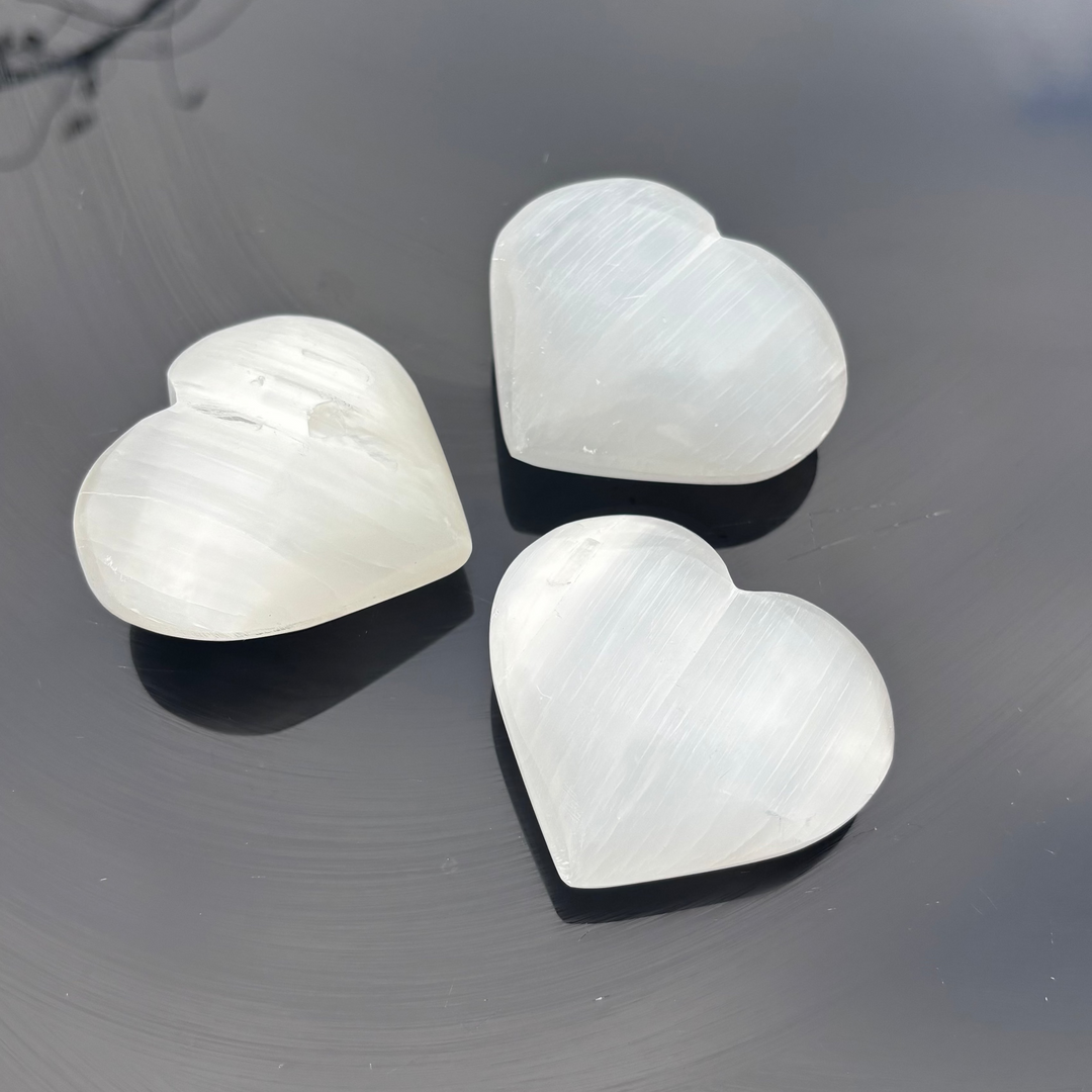 Three polished puffy selenite hearts arranged together on a dark reflective outdoor surface, showing off the stark contrast of their luminous white mineral surfaces, ideal for layout styling on a bedroom nightstand.