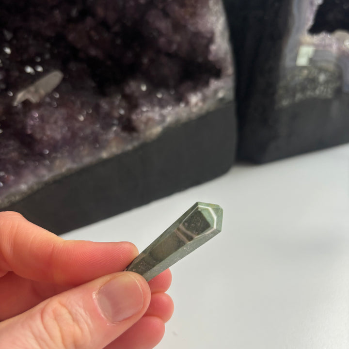 A hand tilting the polished serpentine crystal pocket stone at an angle indoors, detailing the faceted geometric lines and rich olive tones designed to help clear clouded thoughts and bring inner balance.