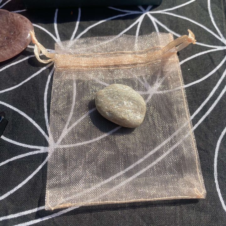 A detailed close-up shot of a sheer tan organza crystal pouch containing a smooth pocket stone. The delicate drawstring mesh packaging rests flat on the sacred geometry fabric, demonstrating how the items are neatly protected within the gift kit.