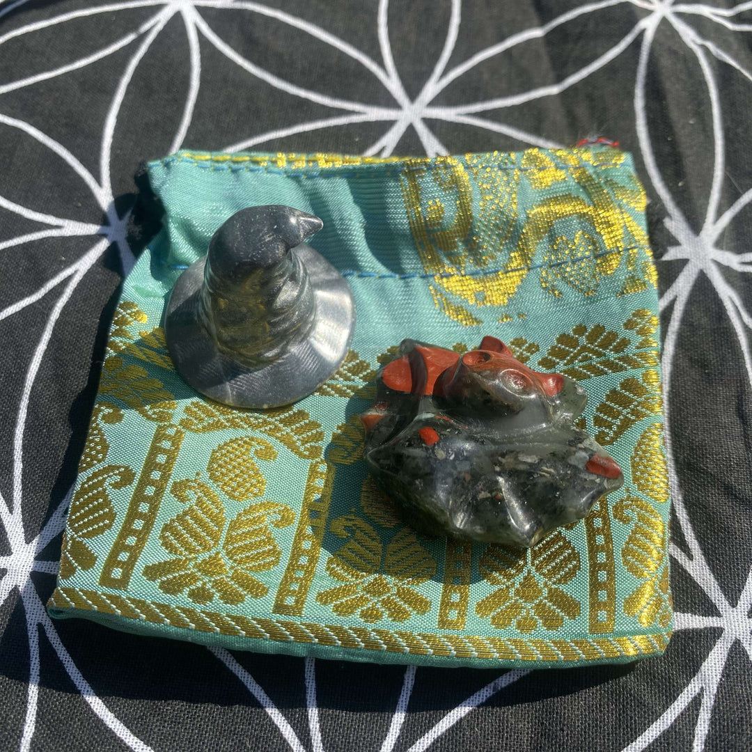 An overhead shot looking down at a carved Silver Obsidian witch's hat and a dark green Bloodstone bat figurine with red speckles. The pair rests flat on an open turquoise and gold jacquard drawstring pouch, showcasing protective root chakra energy against a black and white geometric flower mat.
