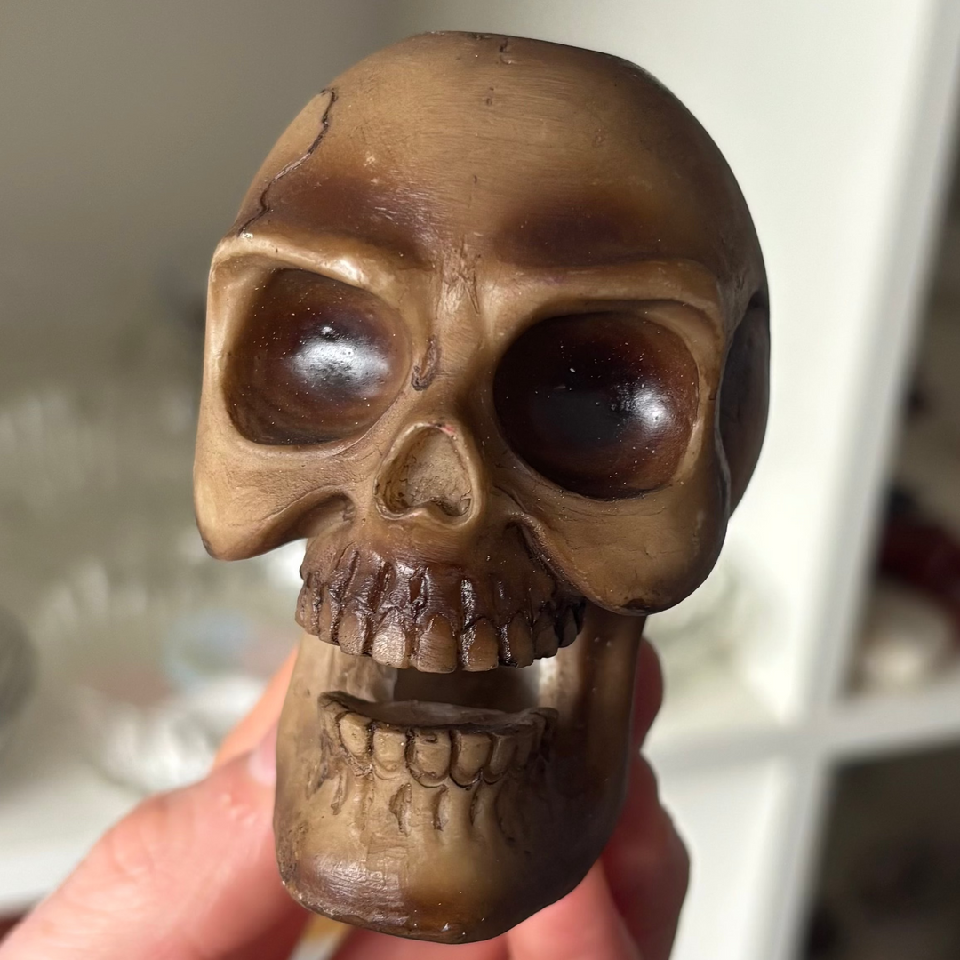 A close-up shot of a hand-carved gothic skull backflow incense burner held gently in a hand. The structured resin body features a weathered metallic bronze wash with realistic bone textures, dark hollow eye sockets, and an open jaw cavity built for holding cascading smoke plumes inside a boutique shop setting.