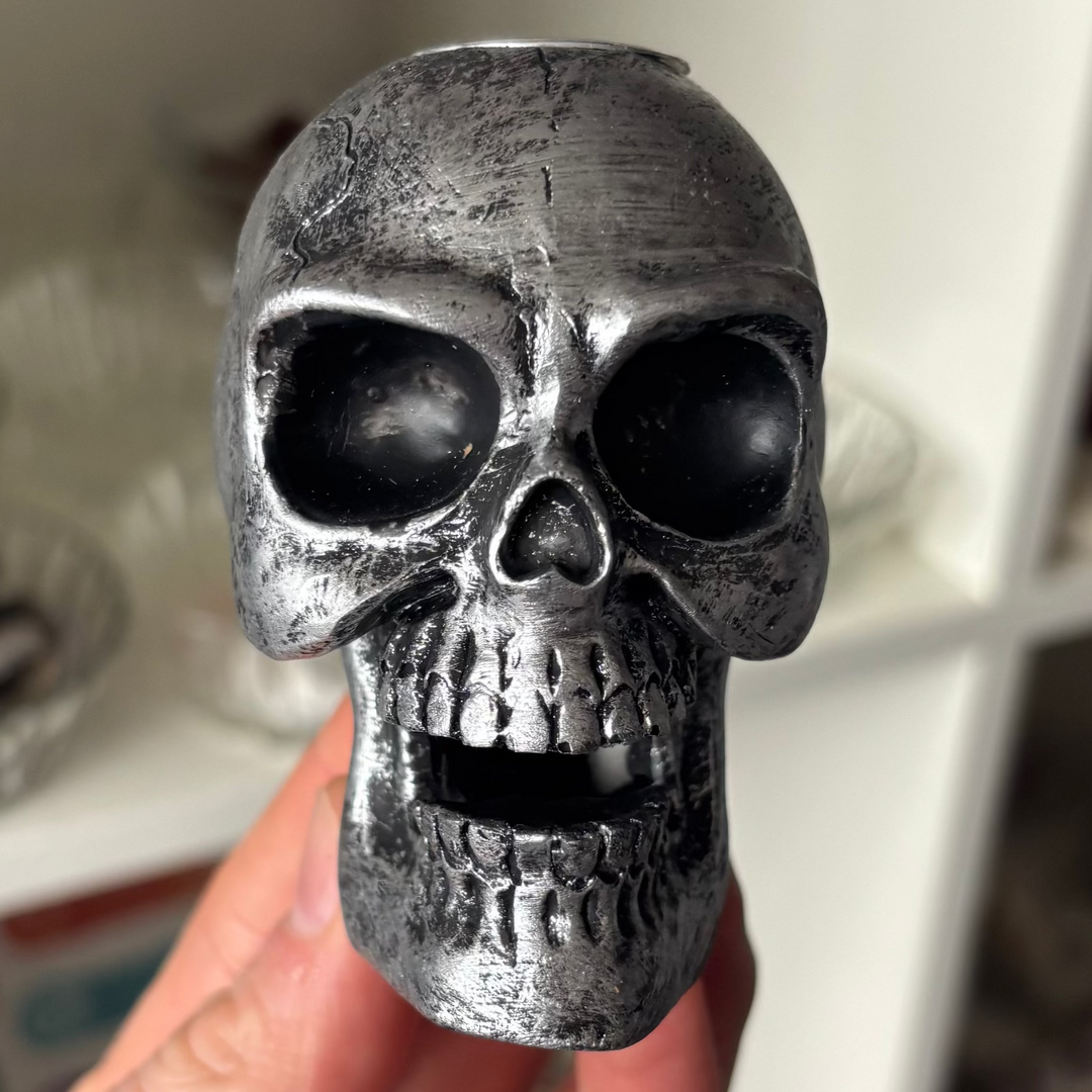 A direct close-up shot of a hand-carved gothic skull backflow incense burner held gently in a hand. The durable resin structure displays a distressed metallic silver finish with realistic bone texturing, hollow black eye sockets, and an open jaw cavity designed for cascading smoke plumes inside a boutique shop setting.