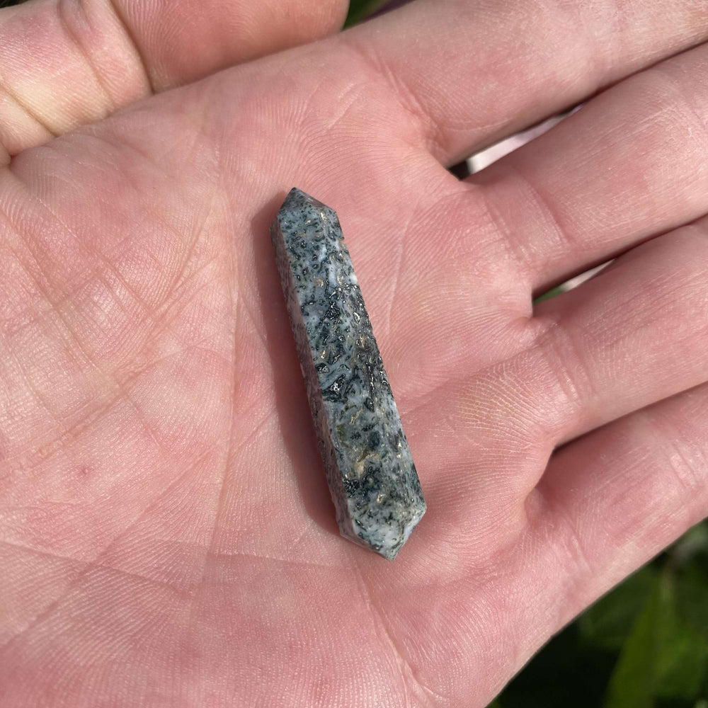 A small, polished Moss Agate double terminated crystal point held vertically between fingers outdoors. The rich green, moss-like inclusions and clear geometric facets stand out beautifully against a blurred background of green foliage and pink garden flowers.