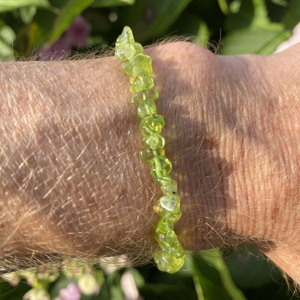 A side-angle outdoor view of the delicate, stretchy crystal bracelet worn on a wrist. The individual, small olive green peridot chips catch the afternoon light, showing off their natural, glassy translucency against a textured background of green garden leaves.