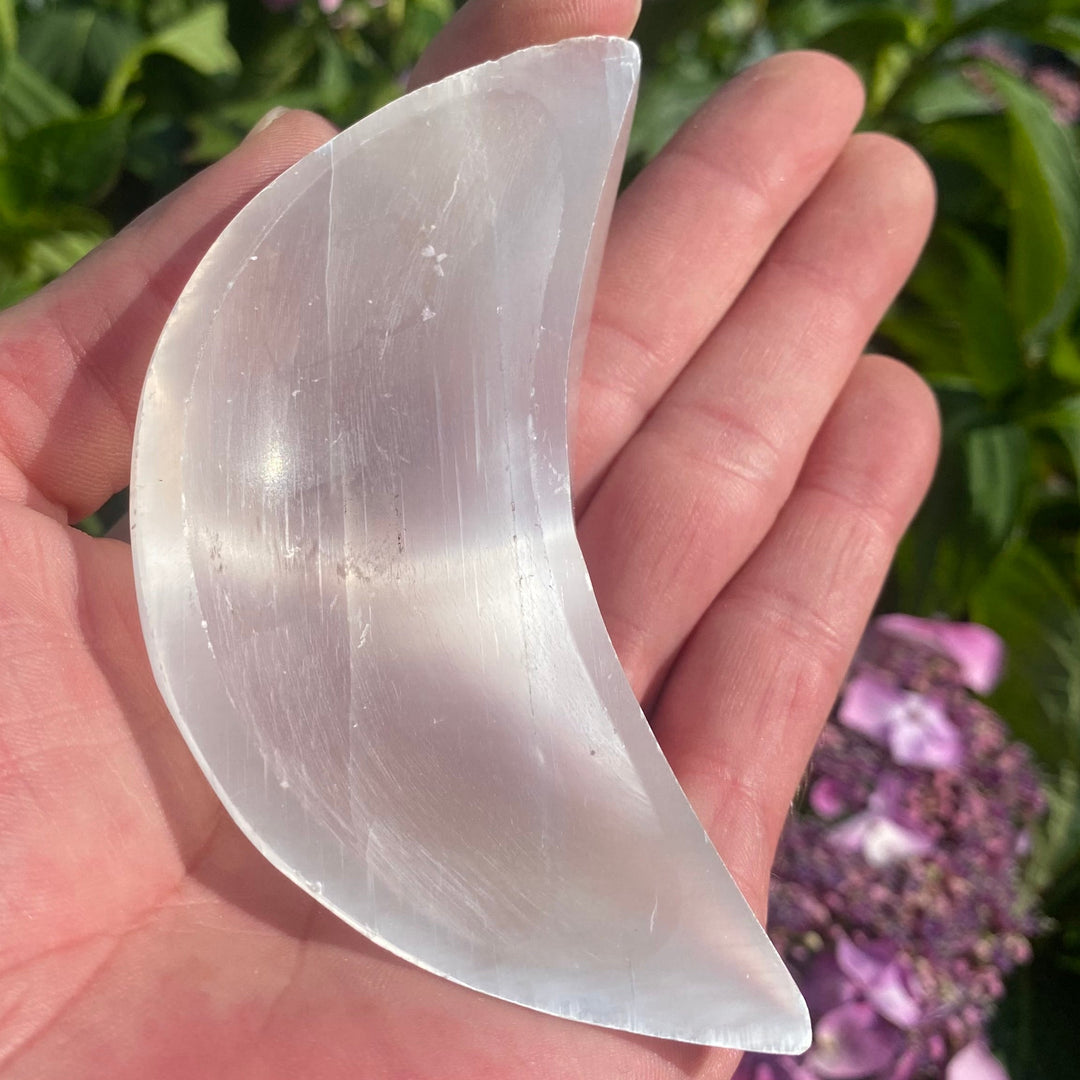 A small polished Selenite moon dish resting in an open palm, showcasing its smooth crescent bowl shape, luminous pearly white surface, and silky mineral grain lines against pink garden flowers.