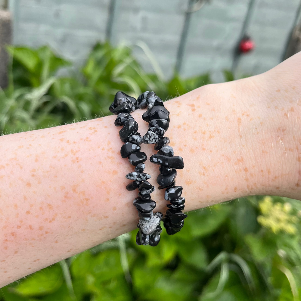 A mid-range view of a dark black gemstone chip bracelet with white snowflake patches worn on an arm against a lush green background