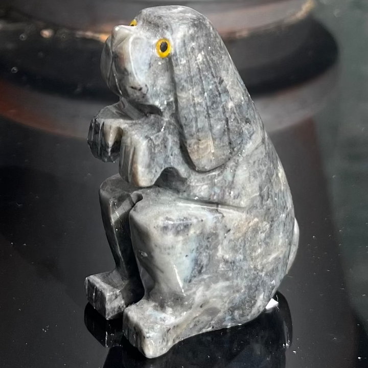 A close-up view of the hand-carved Soapstone Dog Carving sitting on a reflective dark tabletop. The polished pocket-sized animal figurine features a rich, textured grey finish with subtle mineral veins and a tiny golden eye accent detail.