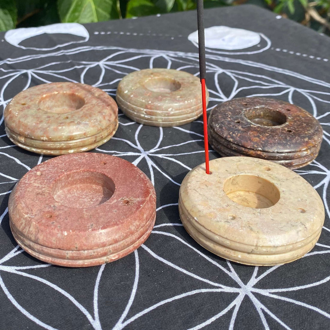 A collection of five circular carved soapstone incense burners in assorted earthy tones arranged outdoors on a black sacred geometry mat. A single black incense stick stands vertically in the front-right beige burner as it rests against a background of green garden leaves.