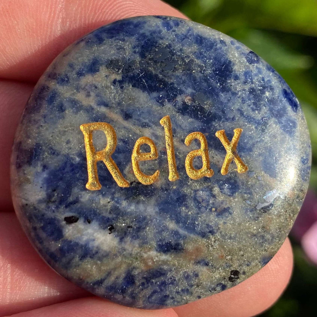 A close-up view of an oval sodalite "RELAX" smoothstone held gently in an open hand. The pocket stone features the word "Relax" engraved in elegant gold lettering on a polished, deep navy blue surface with organic white marbling patterns.