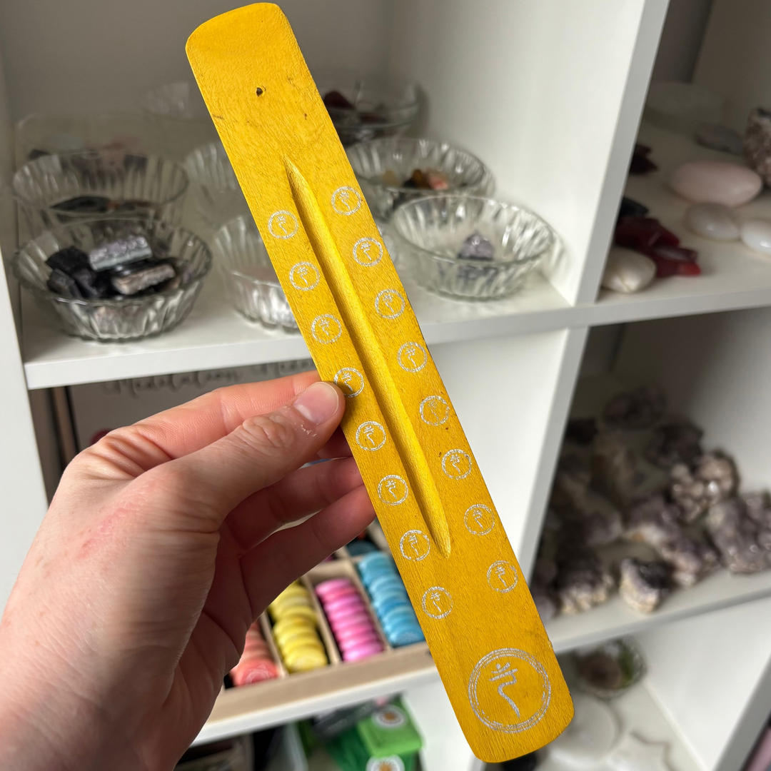 A close-up view of a long, slim wooden incense stick holder held angled in a hand. The smooth wood is finished in a bright sunshine yellow and detailed with a carved Solar Plexus symbol at the base, shown indoors against a white display shelf filled with crystals.
