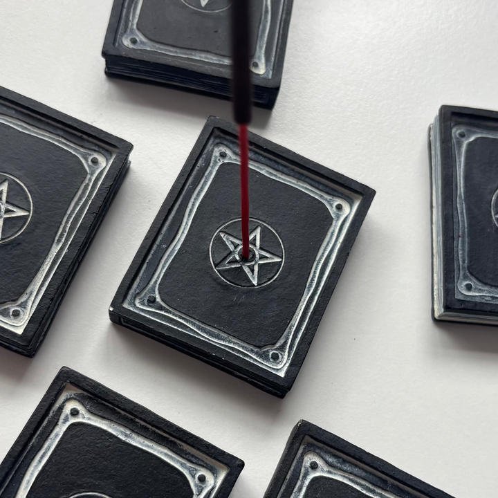 A close-up view of the black resin spell book incense holder resting flat on a white desk with a lit incense stick inserted upright. Aromatic smoke rises gently from the central pentagram hole, demonstrating how this functional ritual accessory creates a cosy sanctuary vibe during meditation pauses.