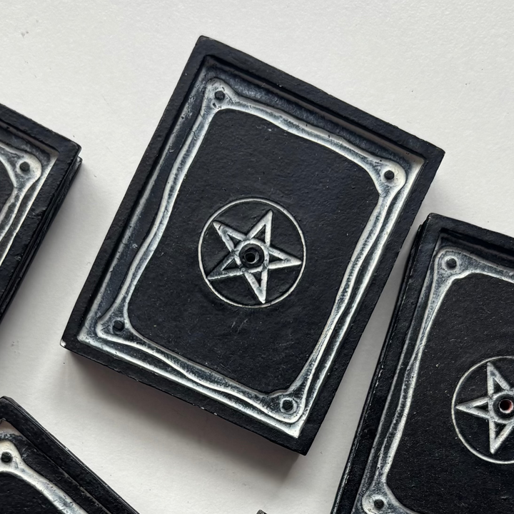 A close-up view of a matte black resin incense stick holder sculpted to look like an open magical spell book grimoire. The detailed plate features a central silver-etched pentagram emblem with a placement hole, bordered by decorative corner scrolls on a clean white table display.
