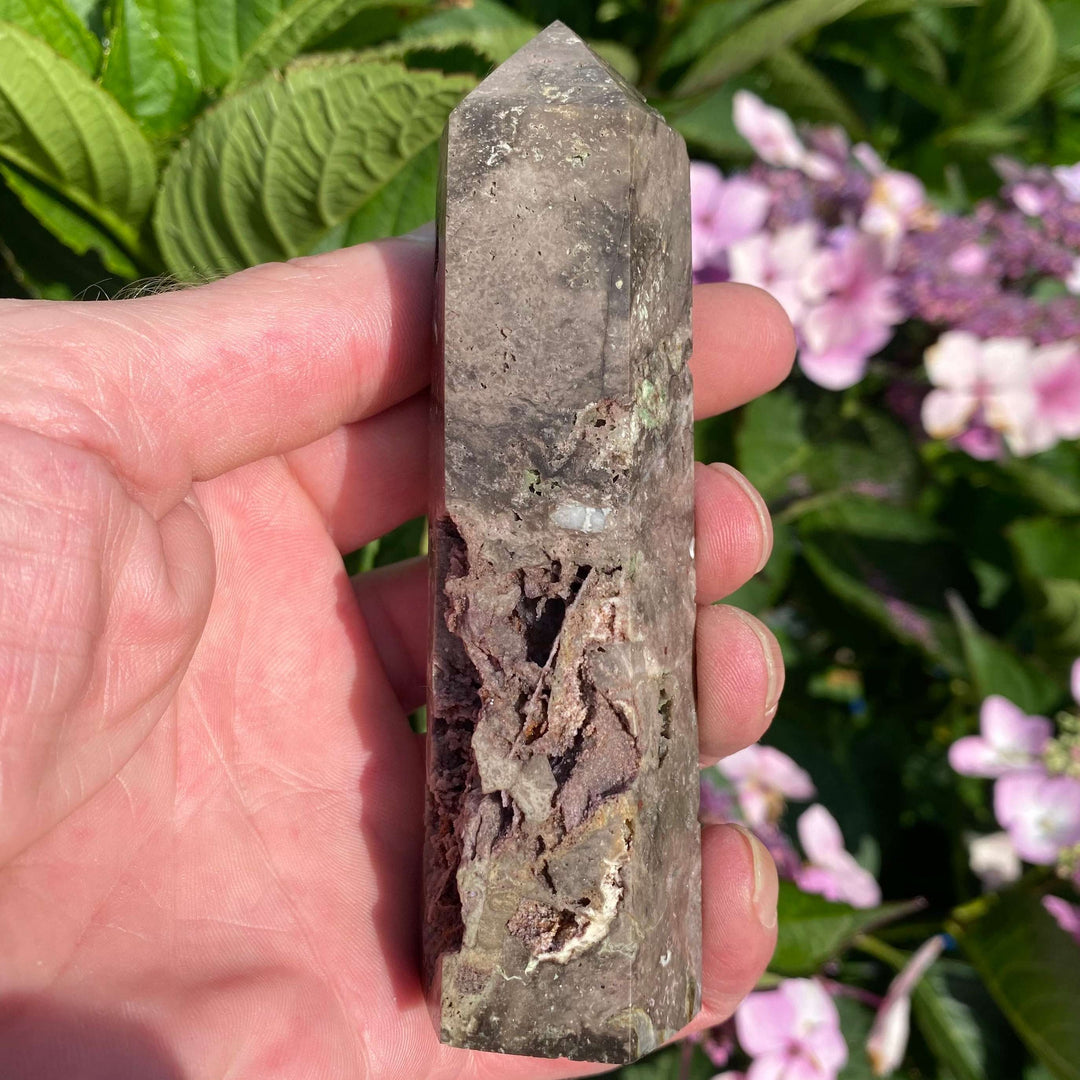 A close-up view of a tall, polished Sphalerite crystal tower held upright in an open hand. The generator obelisk features smooth glassy facets and a dramatic, deep raw cavern pocket on its face, displayed outdoors against a vibrant background of pink garden flowers.