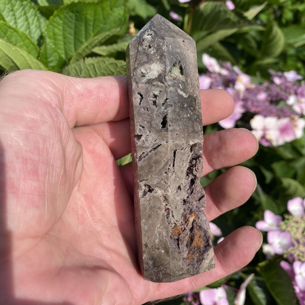 A close-up view of a hand cradling the Sphalerite mineral obelisk outdoors. The natural sunlight highlights its dark charcoal tones, golden druzy flecks, and unique structural markings along its polished sides to create a cosy, comforting vibe for your room.
