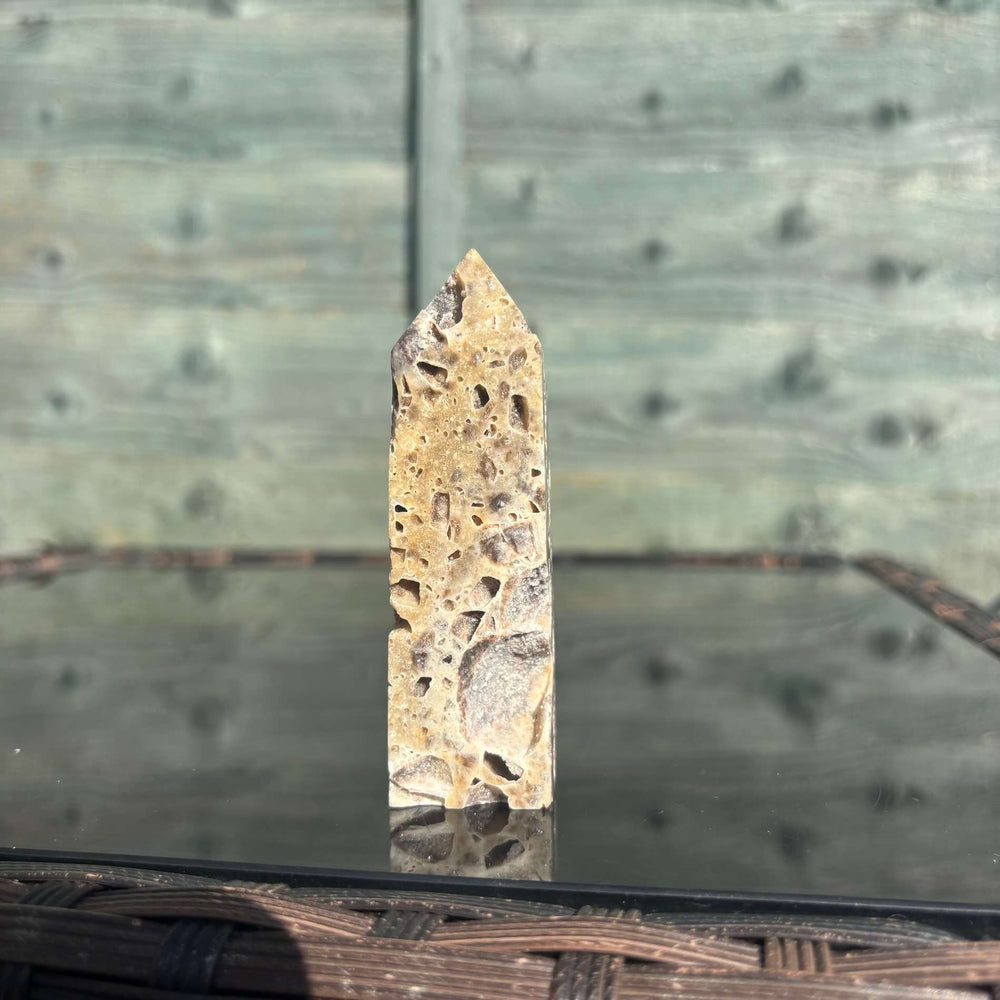 An eye-level shot showing the pointed Sphalerite crystal tower standing alone upon a reflective tabletop display. The outdoor morning sun accentuates its rich earthy patterns and unique geological birthmarks, demonstrating how it adds a steady, protective shield and a grounding presence to any creative workspace.