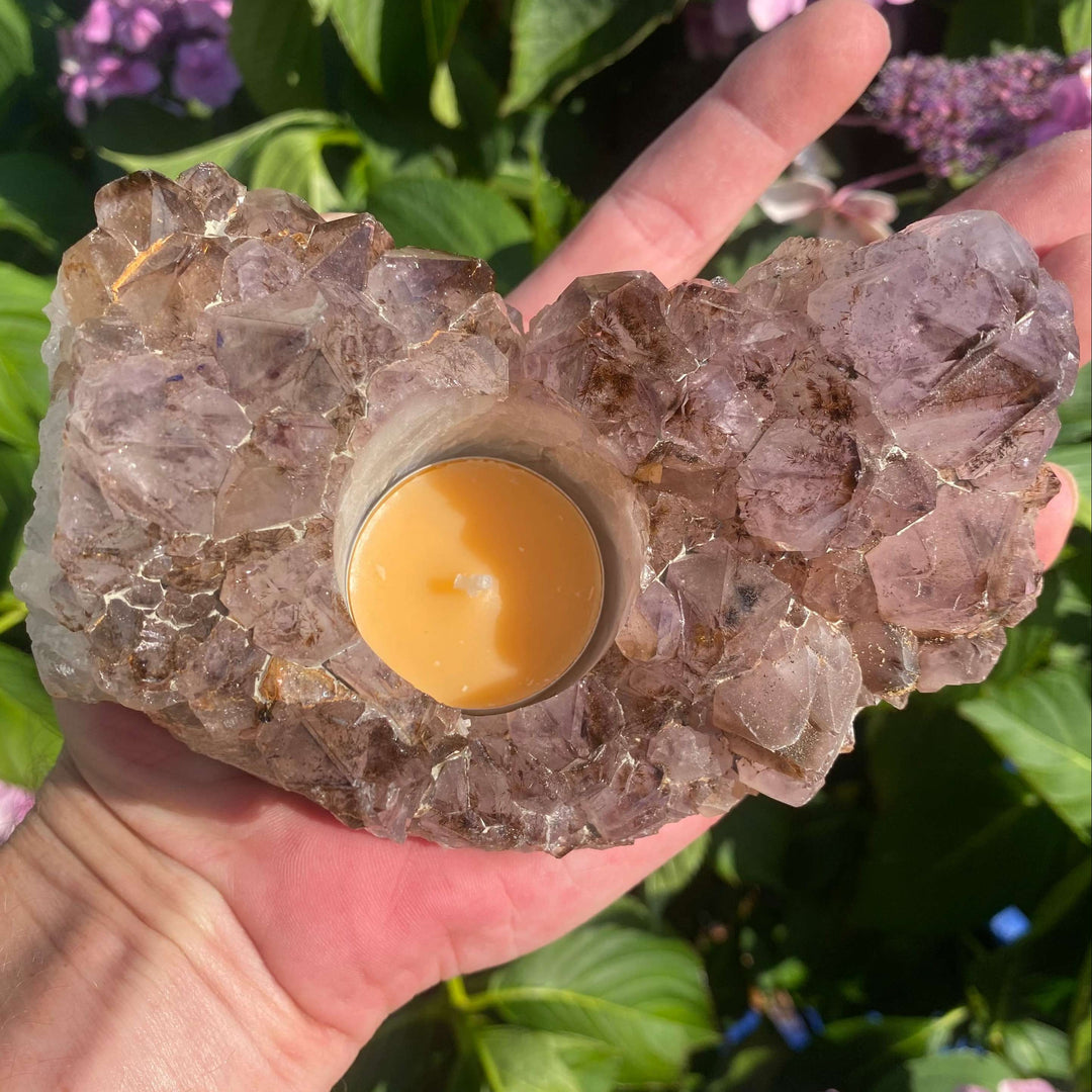 A close-up top-down view of a raw purple Spirit Quartz crystal tea light holder held in an open hand. A yellow wax candle sits inside a custom circular glass cup nested in the center of the glittering, spiky amethyst-hued mineral cluster points.
