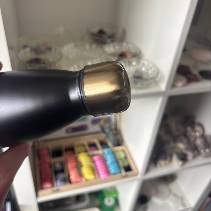 A tilted close-up view highlighting the metallic gold screw-top lid of the black reusable water bottle. This perspective showcases the secure, leak-proof design and twenty-six-centimetre height, making it a reliable companion for a nature walk or workspace desk.
