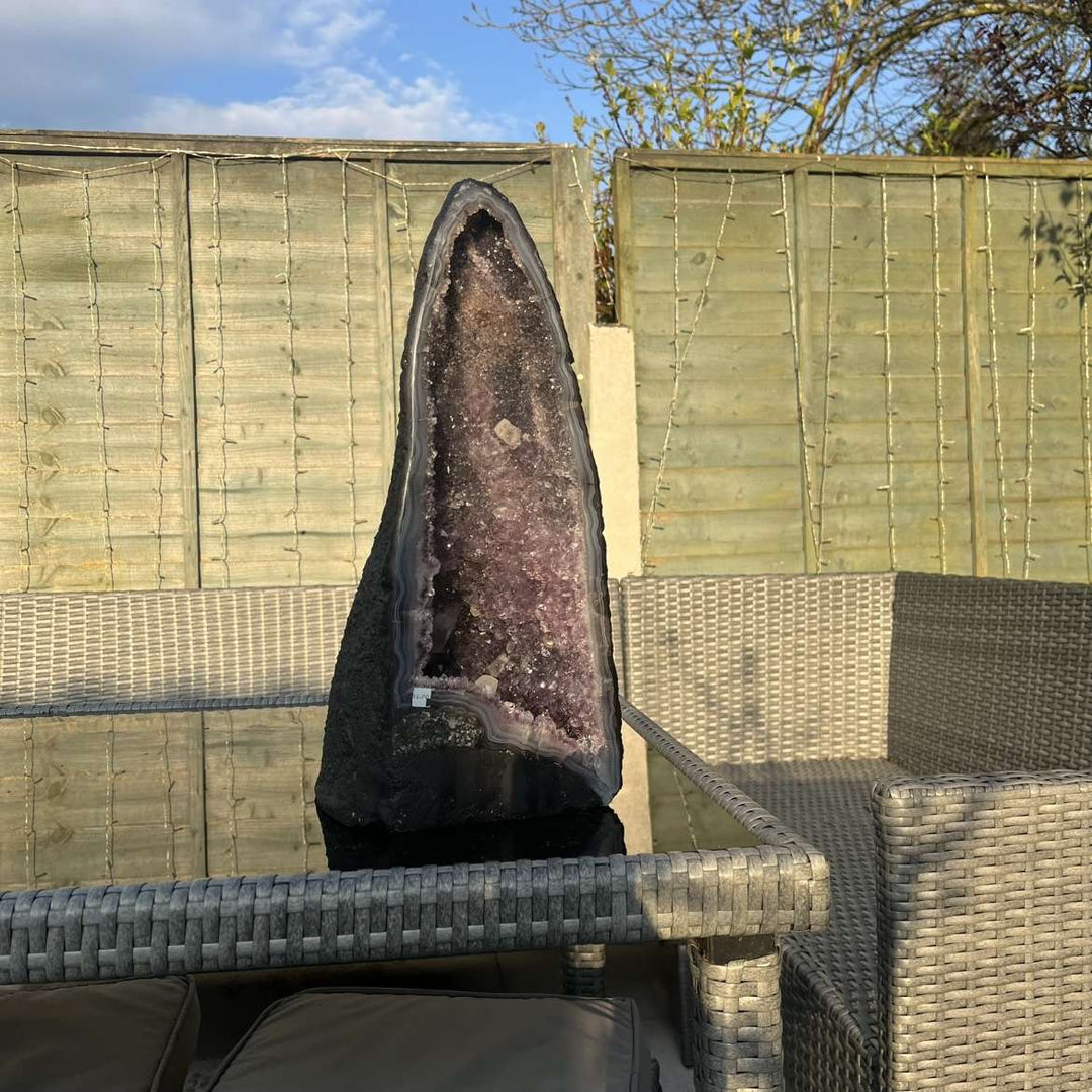A side perspective view of the tall slim amethyst geode standing proudly on an outdoor glass tabletop. The ambient light reflects the fine edges of the stone casing, highlighting a stunning visual anchor to balance crown and third eye centres.  