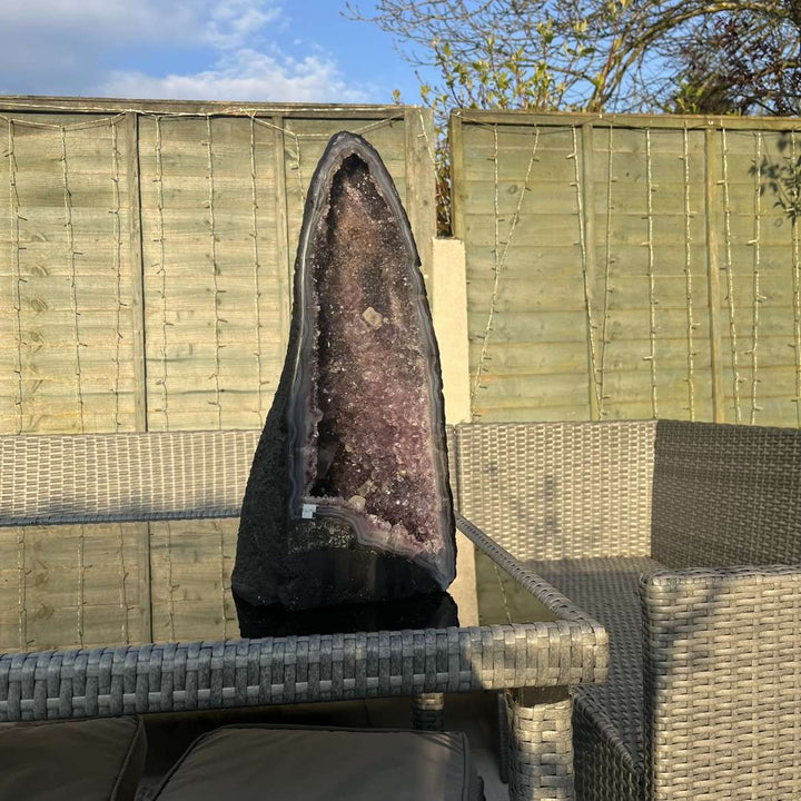 A side perspective view of the tall slim amethyst geode standing proudly on an outdoor glass tabletop. The ambient light reflects the fine edges of the stone casing, highlighting a stunning visual anchor to balance crown and third eye centres.  