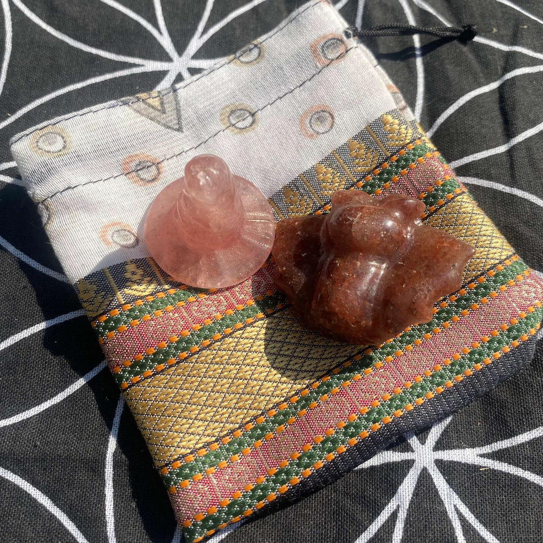 A close-up view of a pink strawberry quartz witch's hat carving and a speckled raspberry aventurine bat figurine resting together on a luxurious gold-patterned white drawstring pouch, styled beautifully against a black and white geometric flower of life fabric backdrop.