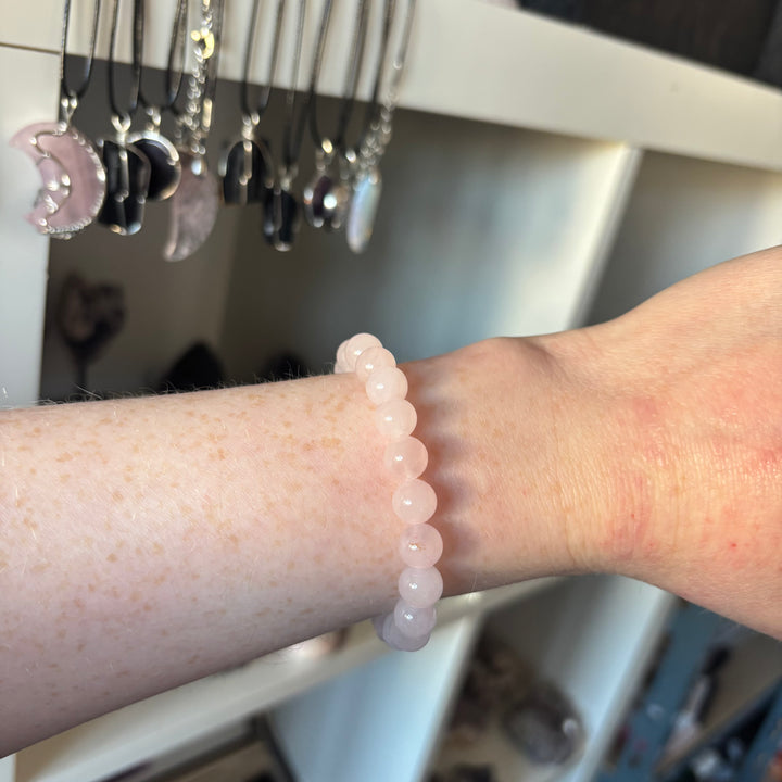 An alternative angled shot of the stretchy Rose Quartz crystal bead bracelet on a wrist, displaying its soft pastel pink tones. The lifestyle perspective highlights its delicate profile inside a crystal shop setting.
