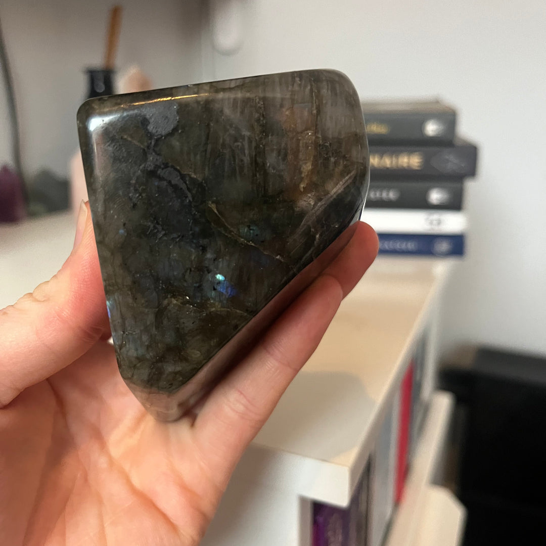 A hand holding the thick side profile of a polished labradorite freeform stone, demonstrating its substantial weight, heavy unpolished base line, and smooth asymmetric curves that protect the room&