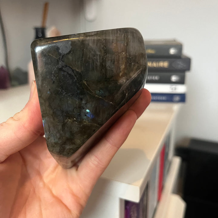 A hand holding the thick side profile of a polished labradorite freeform stone, demonstrating its substantial weight, heavy unpolished base line, and smooth asymmetric curves that protect the room&