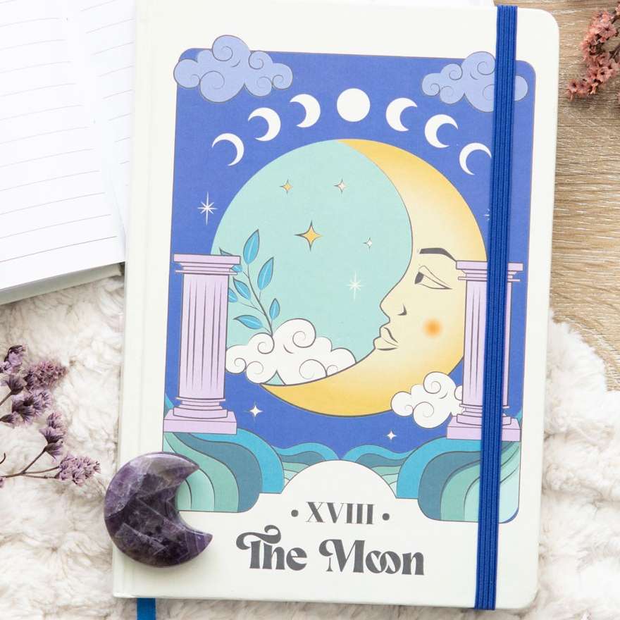 A close-up view of the front cover of The Moon Celestial A5 Notebook resting on a white faux-fur surface next to dried flowers. A small, dark purple lepidolite crystal crescent moon sits on the lower-left corner of the white hardback journal, which features a prominent blue elastic closure band.