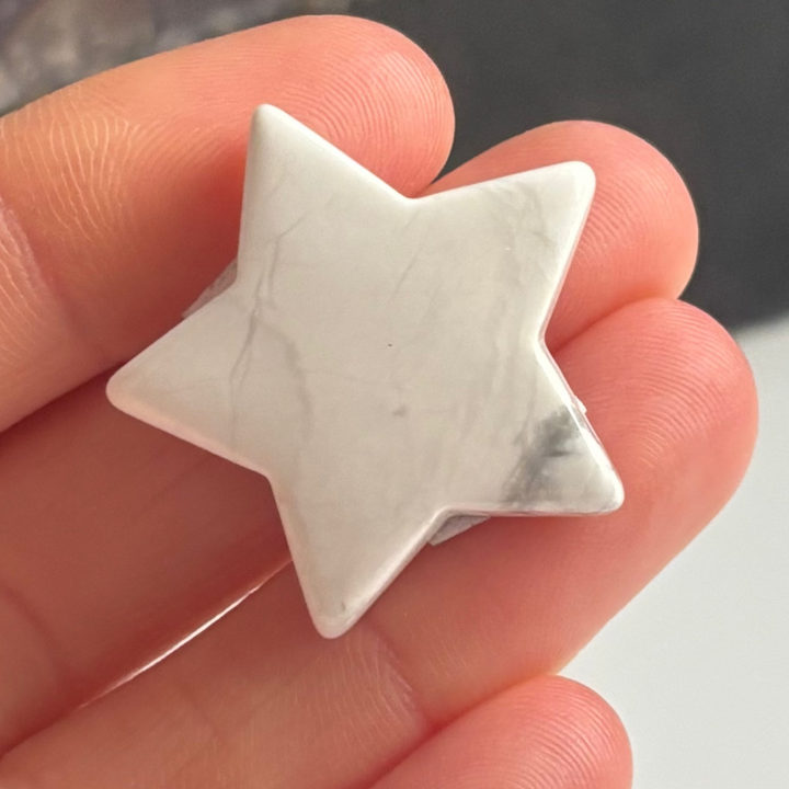 A close-up view of a small polished white howlite star carving held gently across fingertips indoors, detailing its smooth ivory texture, five-pointed silhouette, and soft grey marbling veins for Crown Chakra alignment.
