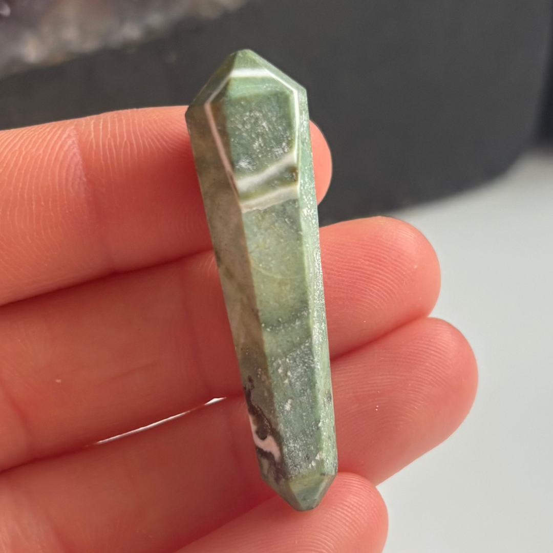 A close-up view of a polished mini serpentine double-terminated point crystal held vertically across fingers indoors, showcasing its unique olive-green colour layers and snake-like skin patterns for Crown Chakra alignment.