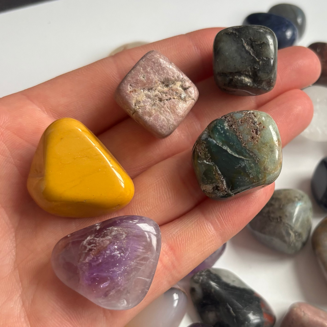 An open hand cradling a selection of polished mystery crystal tumble stones indoors, showcasing a colourful variety including a bright yellow jasper, translucent purple amethyst, and mossy green minerals to show classic pocket sizing scale.