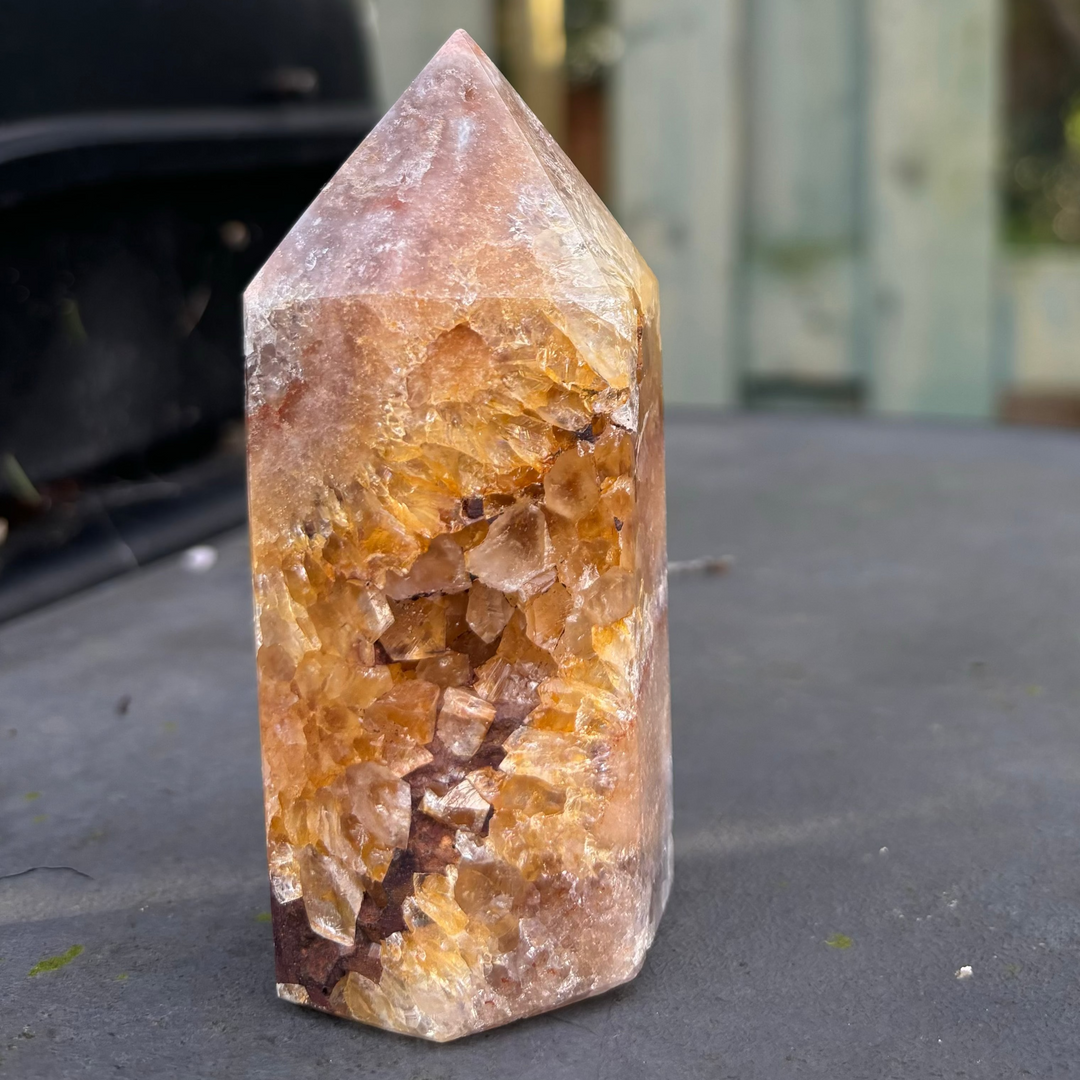 A polished pink amethyst crystal tower standing on a dark surface outdoors, showcasing unique pale rose zones blended with deep golden iron staining and an open crystalline geode pocket for Heart and Crown Chakra alignment.